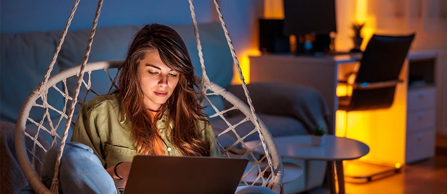 Femeie tânăra care lucrează pe laptop dintr-un balansoar, într-o camera cu lumină ambientală caldă.