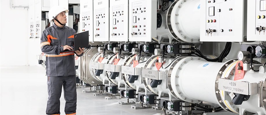Technician inspecting electrical equipment in a substation, using a laptop for monitoring.
