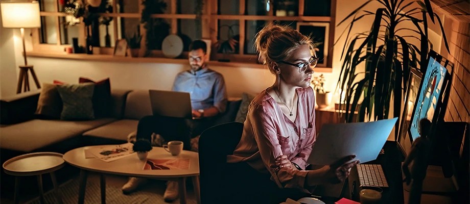 O femeie și un bărbat care lucrează pe computere într-o sufragerie iluminată, pe timp de noapte.